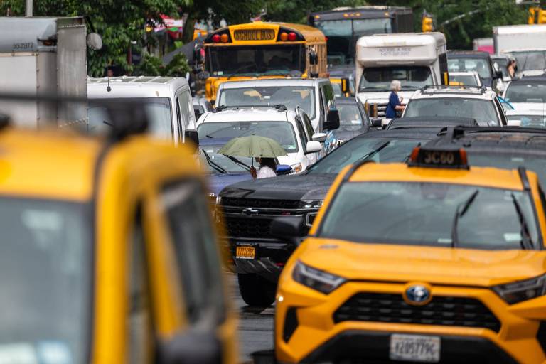 A imagem mostra um congestionamento de veículos em uma rua urbana. Há uma variedade de carros， incluindo um táxi amarelo em primeiro plano e um ônibus escolar ao fundo. Uma pessoa está caminhando com um guarda-chuva， possivelmente para se proteger do sol ou da chuva. O ambiente parece movimentado， com muitos veículos parados e outros em movimento.