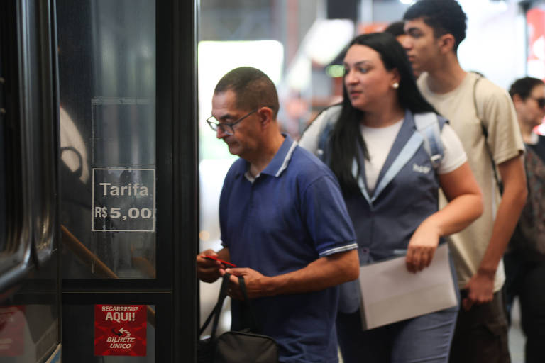 A imagem mostra um grupo de pessoas embarcando em um ônibus. Um homem de camisa azul está na frente， segurando um objeto na mão. Uma mulher com um colete azul e uma folha de papel está logo atrás dele. Ao fundo， outras pessoas também estão na fila para entrar no ônibus. Há um sinal visível na porta do ônibus com o preço da tarifa.