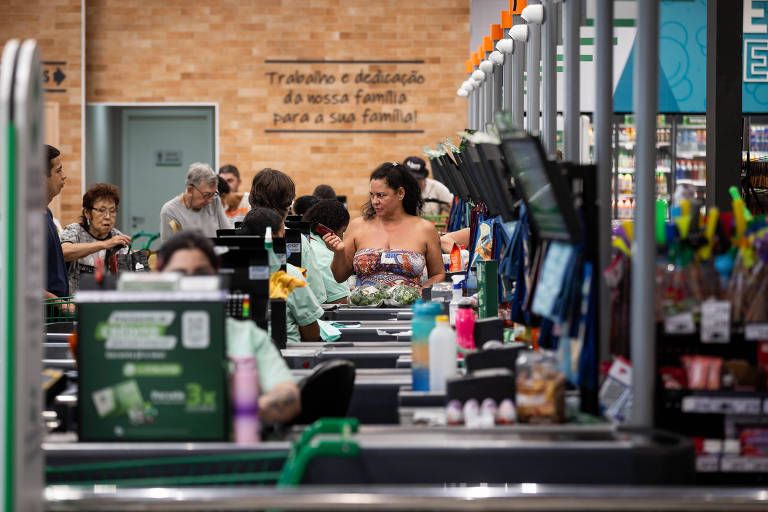 A imagem mostra o interior de um supermercado， com várias pessoas nas filas de caixas. Algumas pessoas estão fazendo compras， enquanto outras estão sendo atendidas. Ao fundo， há uma parede com uma frase em português. O ambiente é bem iluminado e organizado， com produtos expostos nas prateleiras e no balcão dos caixas.