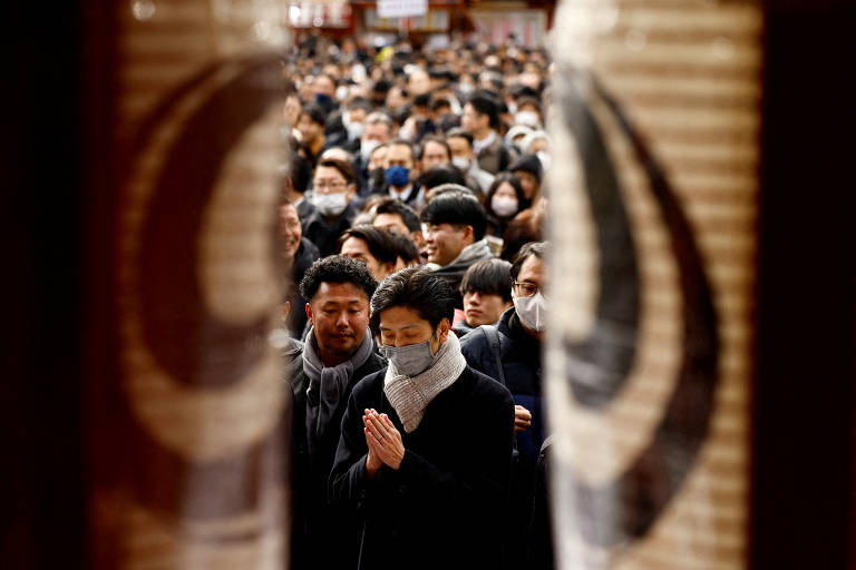 A imagem mostra uma grande multidão de pessoas em um templo, com destaque para um homem em primeiro plano que está de mãos postas em oração. Ele usa uma máscara e um cachecol. Ao fundo, outras pessoas também estão visíveis, algumas com máscaras. A cena é emoldurada por lanternas tradicionais do templo.