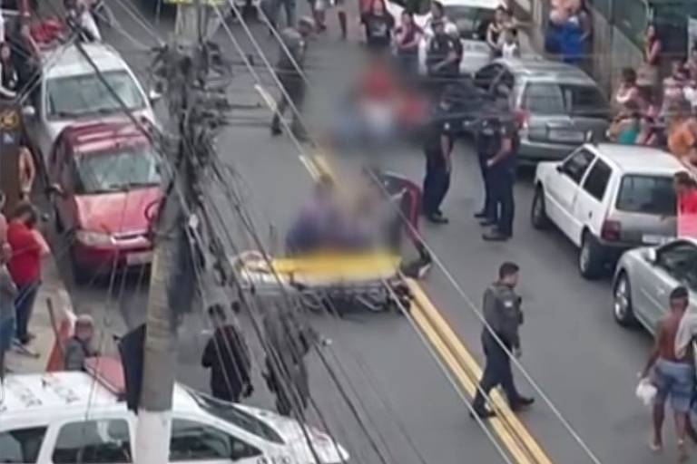 A imagem mostra uma cena em uma rua movimentada， onde várias pessoas estão reunidas. Há um grupo de policiais e uma pessoa sendo atendida em uma maca. Vários veículos estão estacionados ao longo da rua， e há uma multidão observando a situação. Fios elétricos estão visíveis no primeiro plano， e a atmosfera parece tensa.