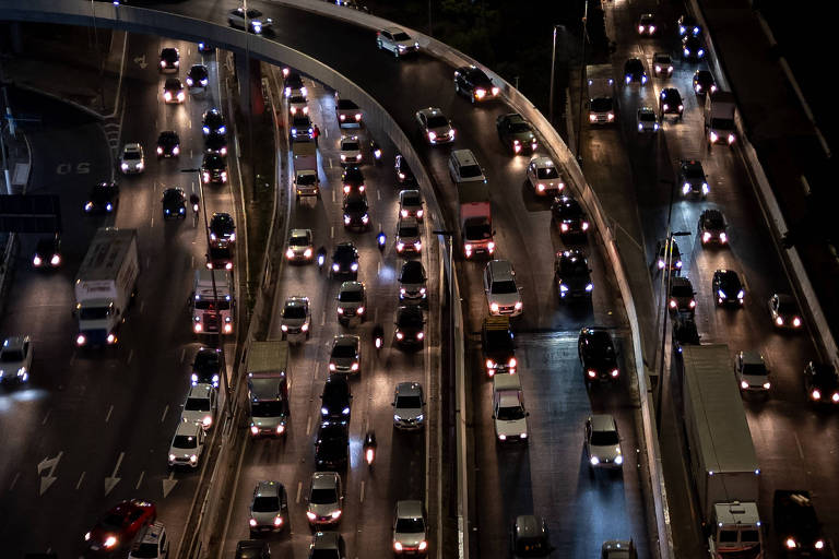 Imagem aérea de uma estrada movimentada à noite， com muitos carros em movimento. As luzes dos veículos iluminam a pista， criando um padrão de luzes brancas e amarelas. A estrada tem várias faixas e curvas， e o tráfego parece intenso.