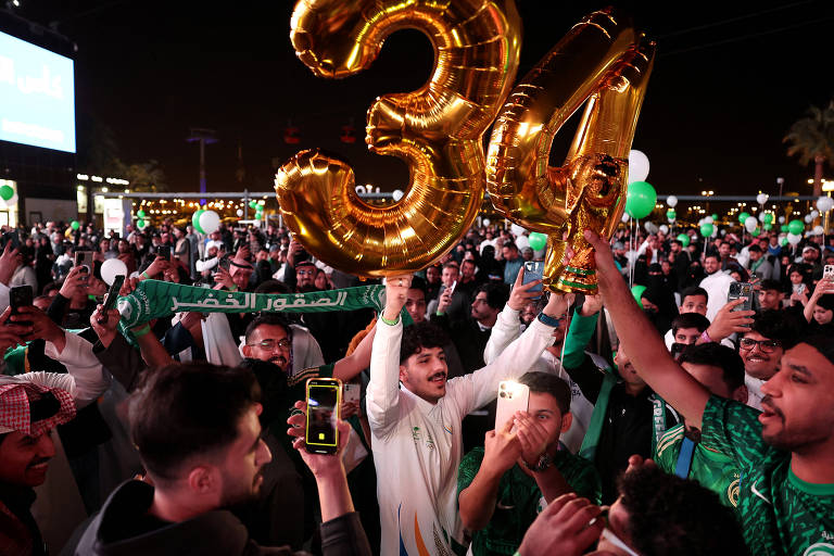 A imagem mostra uma grande multidão celebrando à noite， com pessoas segurando balões dourados em forma dos números 3 e 4. Algumas pessoas estão vestindo roupas tradicionais e outras com camisetas verdes. Há um lenço verde com a inscrição 039;الصقور الخضر039; visível. Muitas pessoas estão usando celulares para registrar o momento， e o ambiente é decorado com balões verdes e brancos.
