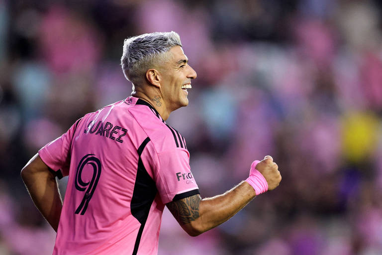 Um jogador de futebol está comemorando， vestindo uma camisa rosa com o nome 039;SUAREZ039; e o número 9 nas costas. Ele tem cabelo platinado e está sorrindo， levantando o punho em sinal de celebração. O fundo da imagem é desfocado， com tons de rosa， sugerindo um ambiente de jogo animado.
