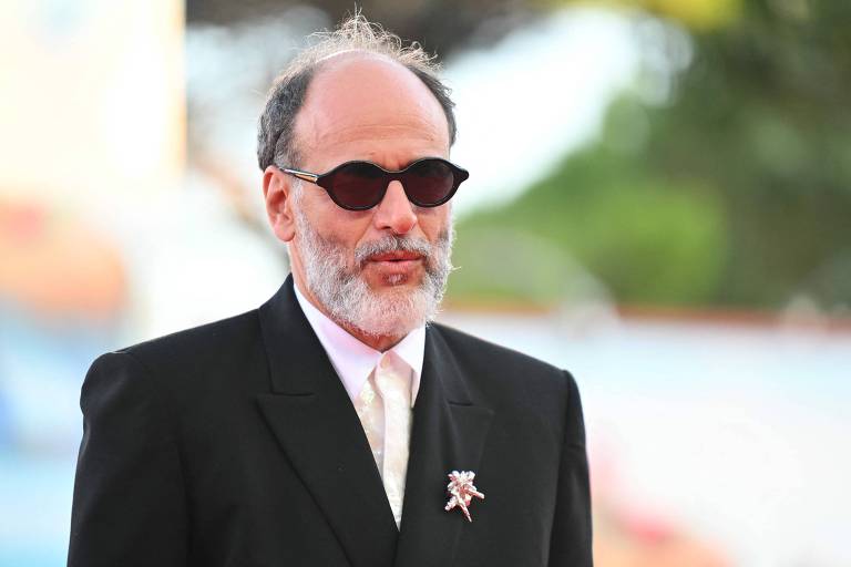 Imagem colorida mostra o diretor Luca Guadagnino. Ele é um homem com barba e cabelo grisalho， usando óculos escuros e um terno preto. Ele está posando em um evento ao ar livre， com um fundo desfocado que sugere um ambiente festivo. Ele também usa uma gravata branca e um broche na lapela do terno.