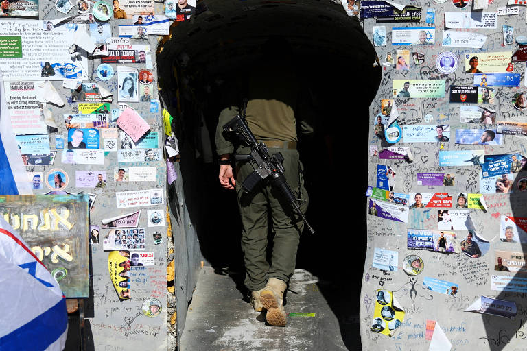 A imagem mostra um soldado caminhando em direção a um túnel escuro， cujas paredes estão cobertas com uma variedade de adesivos e recados. O soldado está vestido com um uniforme militar e carrega uma arma. O ambiente ao redor é colorido devido aos diversos papéis colados nas paredes do túnel