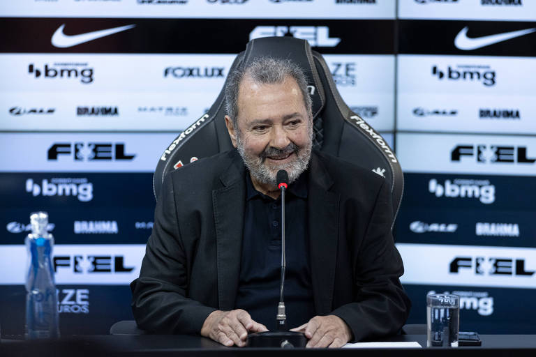 Um homem sorridente está sentado em uma mesa de coletiva de imprensa， com um microfone à sua frente. Ele usa uma camisa preta e um blazer escuro. Ao lado dele， há uma garrafa de água e um copo. O fundo é composto por painéis com logotipos de patrocinadores， incluindo Nike e BMG.
