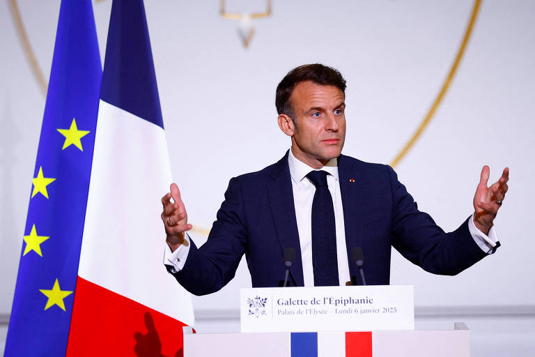 Um homem em um terno escuro está em pé diante de um púlpito， gesticulando com as mãos. Ao fundo， há uma bandeira da França e uma bandeira da União Europeia. O ambiente parece ser uma sala de conferências ou um evento oficial.