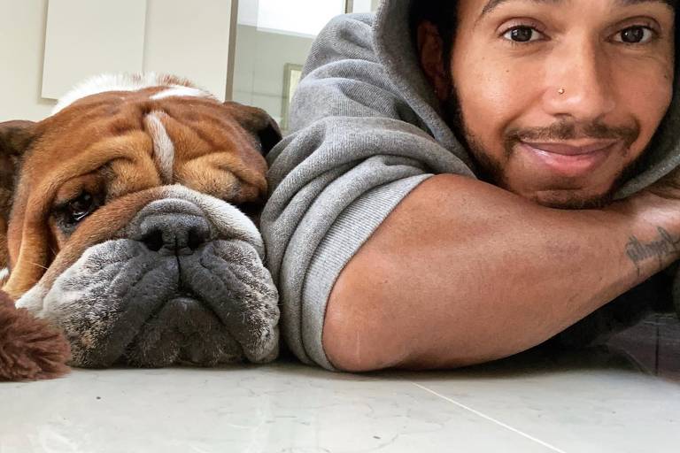 A imagem mostra um homem deitado no chão ao lado de um cachorro da raça bulldog. O homem está usando um moletom cinza e sorri para a câmera, enquanto o cachorro, com pelagem marrom e branca, parece relaxado e apoia a cabeça no chão. Ambos estão em um ambiente interno com piso claro.