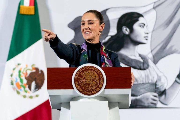 Claudia Sheinbaum discursando em palanque， apontando para frente e com bandeira do México ao fundo
