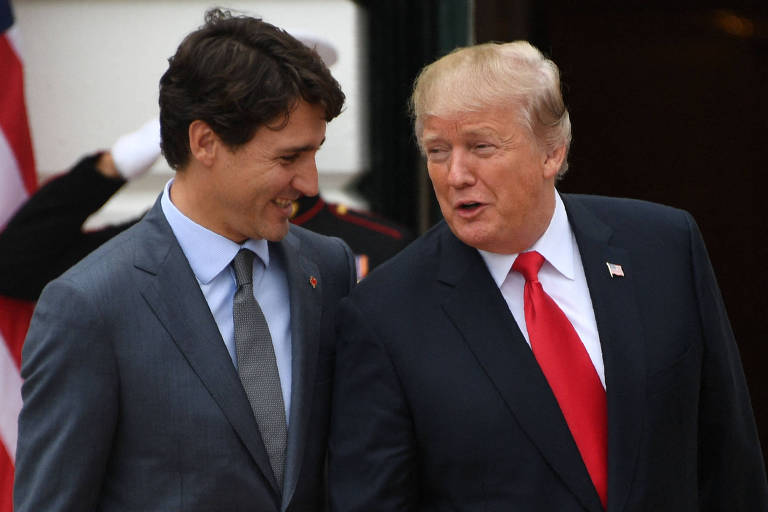 A imagem mostra dois homens conversando em um ambiente externo. Um deles está vestido com um terno cinza e uma camisa clara， enquanto o outro usa um terno escuro com uma gravata vermelha. Ambos parecem sorrir e estão próximos um do outro， com um grupo de pessoas ao fundo， possivelmente em um evento oficial