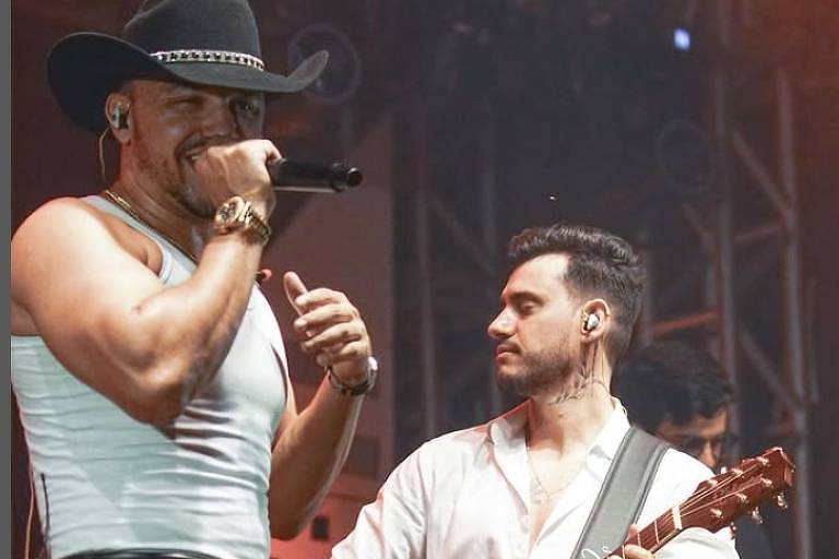 Em foto colorida, dois homens aparecem cantando em cima de um palco