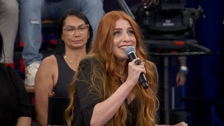 A imagem mostra uma mulher com cabelo longo e ruivo, segurando um microfone e sorrindo enquanto fala. Ela está vestindo uma blusa preta e usa um anel grande. Ao fundo, há uma mulher com cabelo escuro e óculos, observando. A cena parece ser de um programa de televisão, com câmeras visíveis ao fundo.