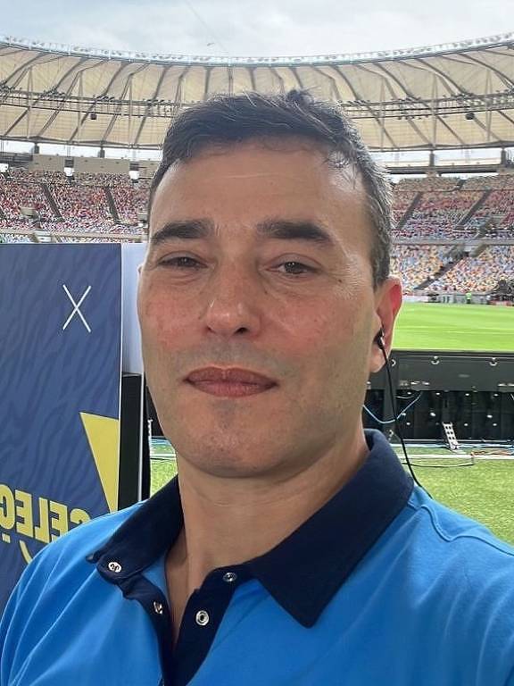 A imagem mostra um homem tirando uma selfie em um estádio. Ele está usando uma camisa azul com gola escura e fones de ouvido. Ao fundo, é possível ver um campo de futebol e uma grande quantidade de torcedores nas arquibancadas. Há também um banner com a palavra 'SELEÇÃO' visível ao lado.