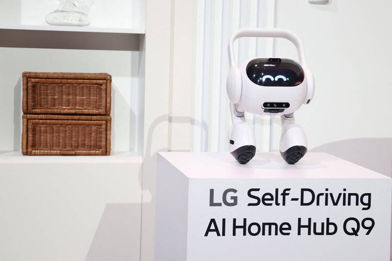 A imagem mostra um robô autônomo da LG， denominado AI Home Hub Q9， posicionado em uma base branca. O robô tem um design amigável， com uma tela que exibe olhos e uma expressão sorridente. Ao fundo， há duas caixas de armazenamento de vime em uma prateleira.