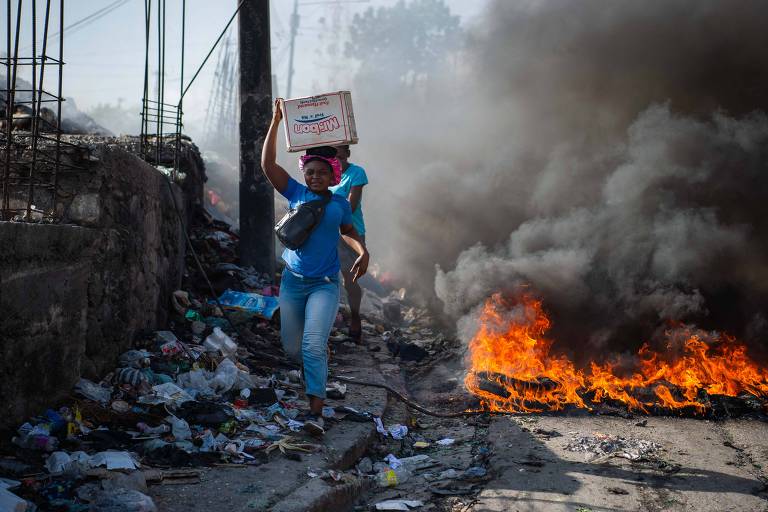 A imagem mostra uma mulher carregando uma caixa sobre a cabeça enquanto caminha em uma rua cheia de lixo. Ao fundo， há fumaça densa e chamas， indicando um incêndio. A mulher está vestida com uma camiseta azul e jeans， e parece estar se afastando da área em chamas.