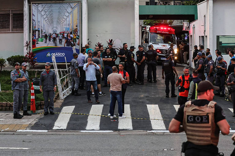 A imagem mostra uma cena de emergência com várias pessoas e policiais em um local. Há uma ambulância visível ao fundo, com luzes piscando. Policiais em uniformes estão posicionados em diferentes áreas, enquanto algumas pessoas, incluindo civis, estão na calçada. Um grande cartaz com imagens de pessoas em um ambiente interno é visível na parede ao fundo.