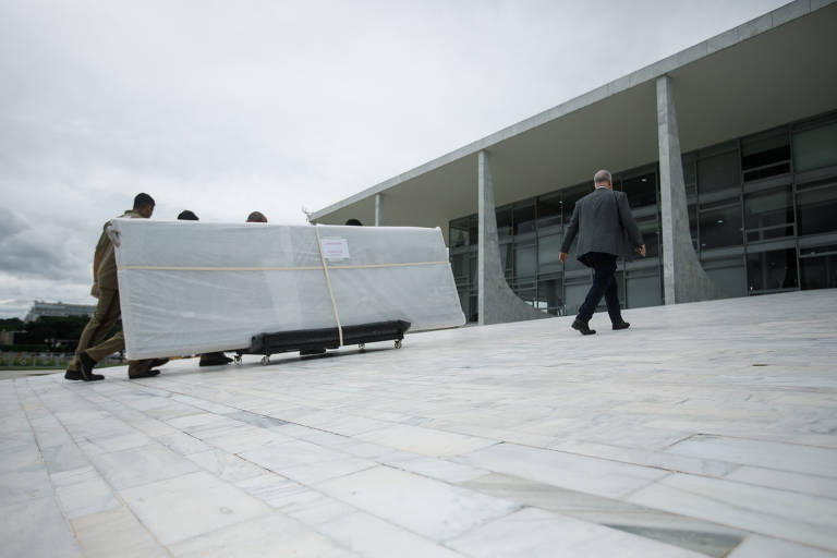 Na imagem, um grupo de pessoas está transportando um objeto grande e coberto com uma lona branca. Eles estão se movendo em direção a um edifício moderno, que possui grandes janelas de vidro. Um homem de terno está caminhando em direção ao edifício, enquanto os outros carregam o objeto. O céu está nublado, e o piso é de pedras claras.