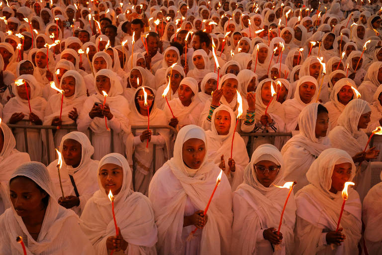 A imagem mostra uma grande multidão de pessoas vestidas com roupas brancas, segurando tochas acesas. As pessoas estão em um ambiente ao ar livre, com a maioria usando lenços na cabeça. A cena é iluminada pelas chamas das tochas, criando um efeito de luz quente. A multidão parece estar unida em um evento ou celebração.