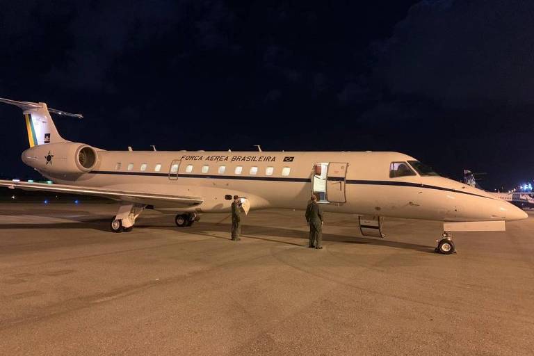 A imagem mostra um avião executivo estacionado em uma pista durante a noite. O avião é de cor clara， com detalhes em azul e dourado. Duas pessoas estão em pé ao lado do avião， e o ambiente ao redor é escuro， com algumas nuvens visíveis no céu.