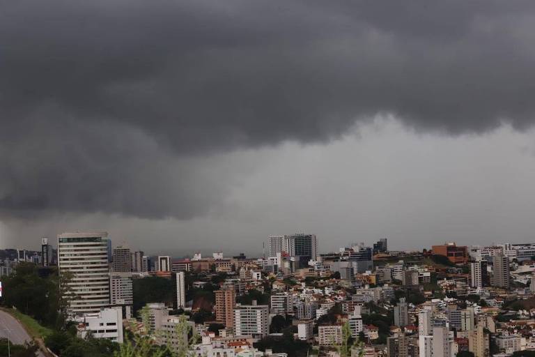 A imagem mostra uma vista panorâmica de uma cidade sob um céu nublado e escuro. As nuvens são densas e ameaçadoras， cobrindo a maior parte do céu. A cidade é composta por diversos edifícios， com uma mistura de estruturas altas e baixas， e algumas áreas verdes visíveis entre os prédios. A iluminação é suave， sugerindo que pode estar prestes a chover.