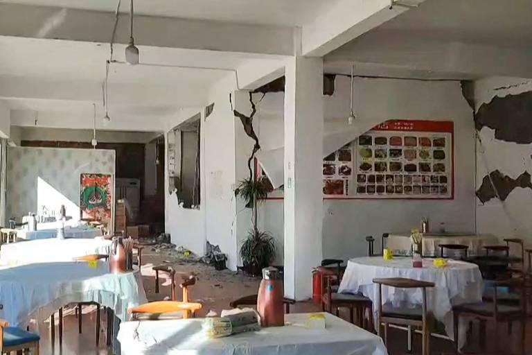 A imagem mostra o interior de um restaurante com paredes danificadas, apresentando rachaduras visíveis. Mesas com toalhas brancas estão dispostas pelo espaço, algumas com garrafas e utensílios. O chão está bagunçado, com detritos e objetos espalhados. Uma parede exibe um quadro com imagens de pratos, enquanto a iluminação é fornecida por lâmpadas penduradas no teto.