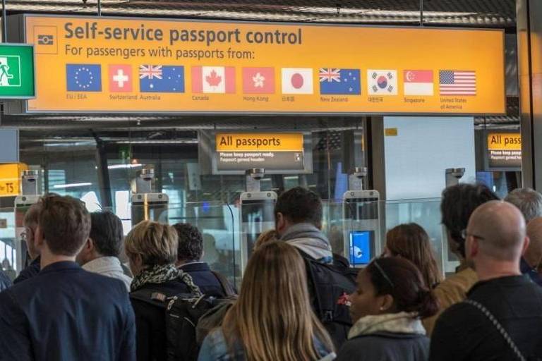 A imagem mostra uma fila de pessoas em um controle de passaporte self-service em um aeroporto. Acima， há um painel informativo com o título 039;Self-service passport control039; e a frase 039;For passengers with passports from:039; seguida de bandeiras de vários países， incluindo a União Europeia， Reino Unido， Canadá， Japão， Austrália， Coreia do Sul e Estados Unidos. O ambiente é moderno e bem iluminado.