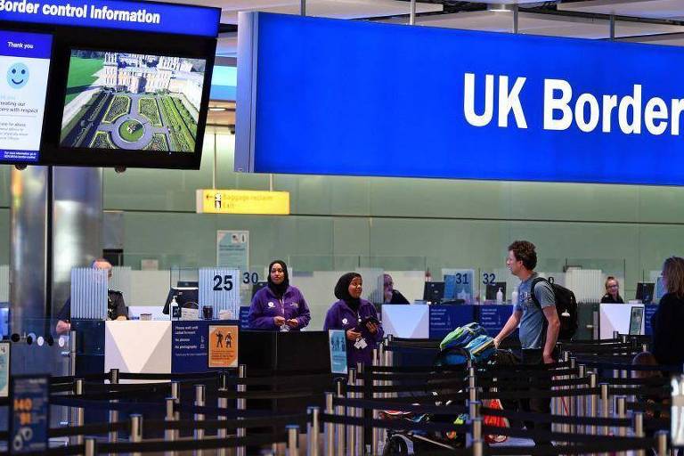 A imagem mostra a área de controle de fronteira do Reino Unido em um aeroporto. Há um grande sinal azul com a inscrição 039;UK Border039; e um painel eletrônico exibindo informações sobre o controle de fronteira. Algumas pessoas estão na fila， enquanto funcionários em uniformes roxos estão presentes. O ambiente é moderno e bem iluminado.