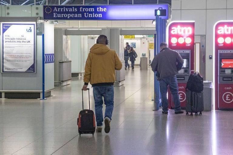 A imagem mostra um terminal de aeroporto com sinalização indicando 039;Chegadas da União Europeia039;. Um homem com uma mala de rodinhas está caminhando em direção à área de chegadas， enquanto outros passageiros estão próximos a caixas eletrônicos. O ambiente é moderno e bem iluminado.