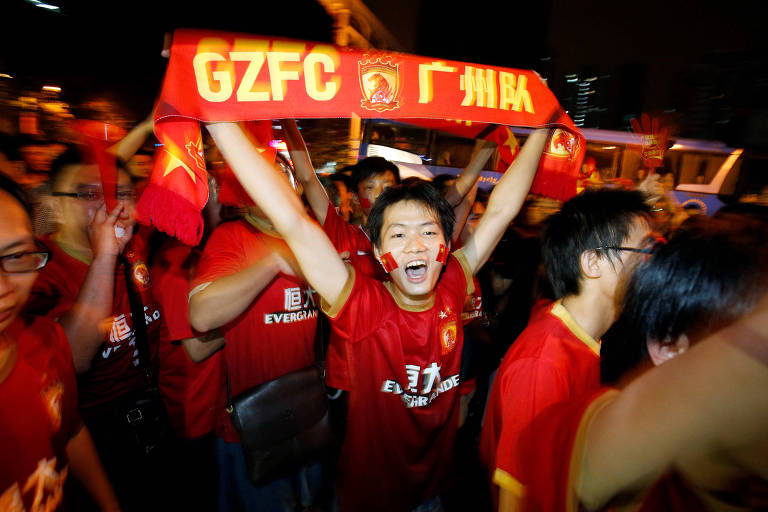 Um grupo de torcedores está celebrando à noite， todos vestindo camisetas vermelhas. Um jovem no centro levanta um cachecol vermelho com as letras 039;GZFC039; e grita de alegria. Outros torcedores ao redor também estão animados， alguns com bandeirinhas e pinturas no rosto. O ambiente é festivo， com luzes ao fundo e uma atmosfera de celebração.
