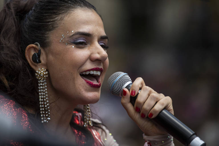 Uma mulher canta em um microfone, sorrindo e com expressão animada. Ela usa brincos grandes e tem maquiagem brilhante. Seu cabelo está preso em um coque, e suas unhas estão pintadas de vermelho.