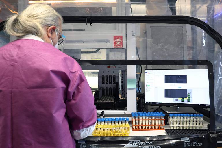 A imagem mostra um ambiente laboratorial onde uma pessoa， com jaleco ou roupa de proteção na cor rosa， está trabalhando em frente a uma bancada equipada com equipamentos laboratoriais. Há várias amostras em tubos organizados em racks coloridos sobre a bancada， indicando o preparo ou análise de materiais. Ao fundo， há uma estrutura de vidro ou acrílico， possivelmente uma cabine de fluxo laminar， e duas telas de computador exibindo dados ou imagens relacionados ao processo em andamento. 





