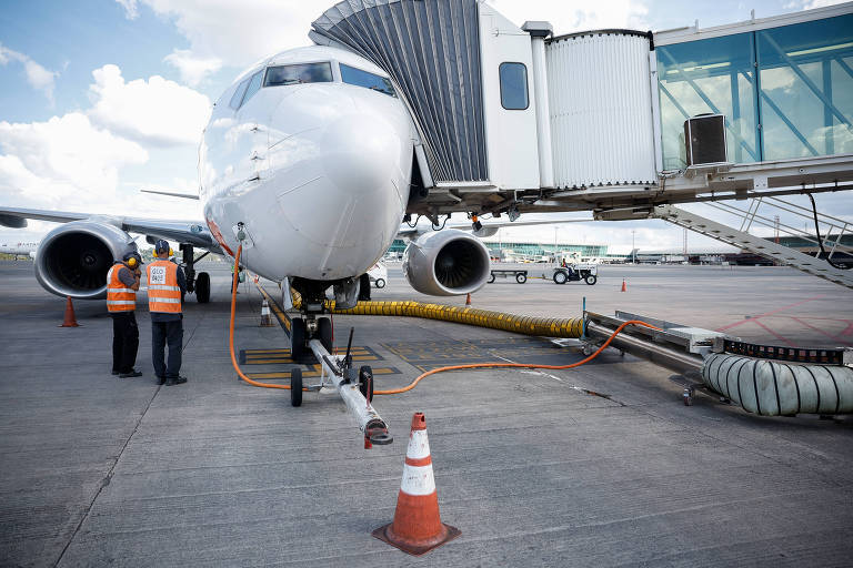 A imagem mostra um avião estacionado em um terminal de aeroporto， com uma ponte de embarque conectada. Dois funcionários， usando coletes laranja， estão próximos ao avião， que possui um tubo laranja conectado a ele. Há cones de sinalização no chão e um edifício de terminal ao fundo sob um céu parcialmente nublado.