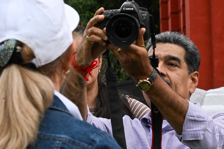 Um homem está segurando uma câmera Canon e tirando uma foto de uma mulher que está de costas. A mulher usa um boné branco e uma jaqueta jeans. Ao fundo， há outras pessoas e uma estrutura vermelha.