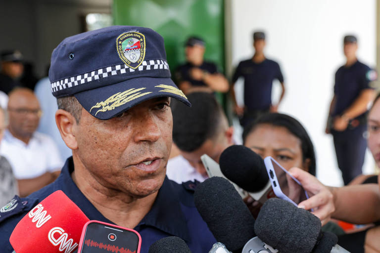 Um oficial de segurança， usando um uniforme e um boné com insígnias， está falando para a imprensa. Ele é cercado por repórteres com microfones， enquanto ao fundo， outras pessoas em uniformes estão visíveis. O ambiente parece ser uma área pública.