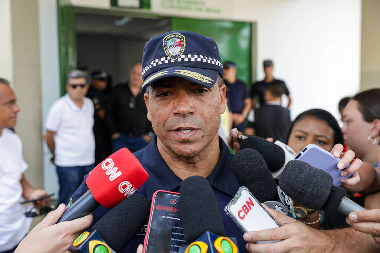 Um homem com uniforme de policial e boné está falando para a imprensa， cercado por microfones de diferentes veículos de comunicação. Ao fundo， há pessoas observando a coletiva. O ambiente parece ser uma área pública， possivelmente uma delegacia ou centro de segurança.