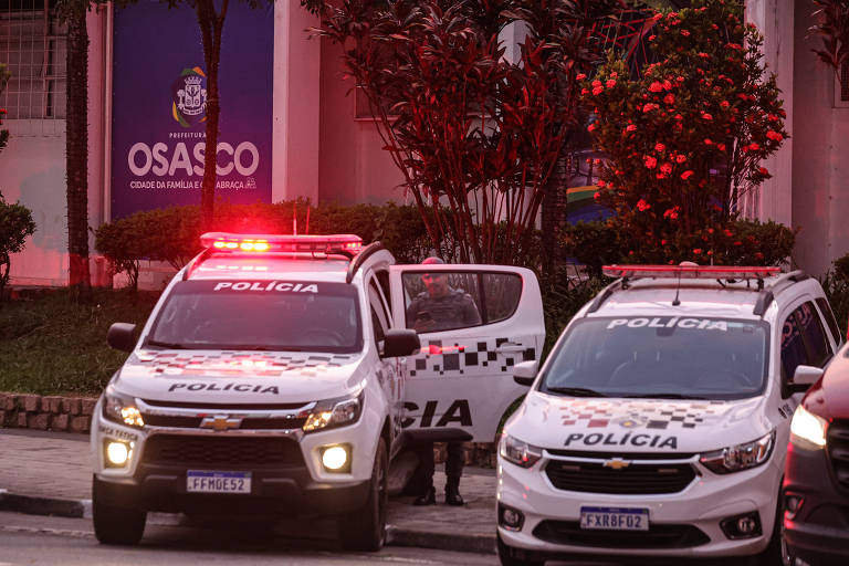 A imagem mostra dois veículos policiais estacionados em uma rua. Ambos os carros têm luzes de emergência vermelhas acesas. Ao fundo， há um edifício com uma placa que diz 039;OSASCO039;. A cena é iluminada por uma luz suave do ambiente， e há vegetação ao redor.