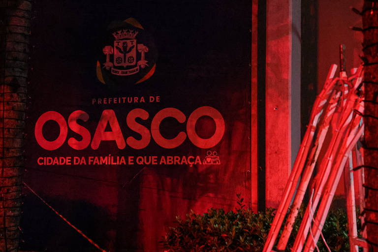 A imagem mostra a fachada da Prefeitura de Osasco iluminada em vermelho. O logotipo da prefeitura está visível no topo, seguido pelo texto 'PREFEITURA DE OSASCO' e a frase 'CIDADE DA FAMÍLIA E QUE ABRAÇA'. Ao lado, há um conjunto de objetos brancos, possivelmente tubos ou bastões, apoiados em uma parede.