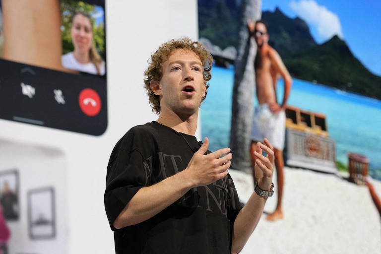 Imagem mostra Mark Zuckerberg， um homem com cabelo cacheado e uma camiseta preta está falando em um palco. Ao fundo， há uma tela mostrando uma imagem de uma praia com montanhas e uma pessoa ao lado de um tronco de árvore. O homem gesticula com as mãos enquanto se dirige ao público.