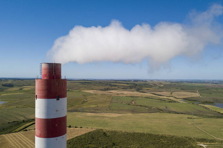 A imagem mostra uma chaminé de uma usina, pintada com listras vermelhas e brancas, emitindo uma nuvem de fumaça branca. Ao fundo, há uma vasta área de campos verdes e um céu azul claro.