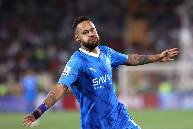 Um jogador de futebol está em campo， vestindo um uniforme azul com detalhes brancos. Ele está fazendo um gesto de celebração， com os braços abertos e uma expressão facial de satisfação. O fundo mostra uma multidão desfocada， indicando um evento esportivo.
