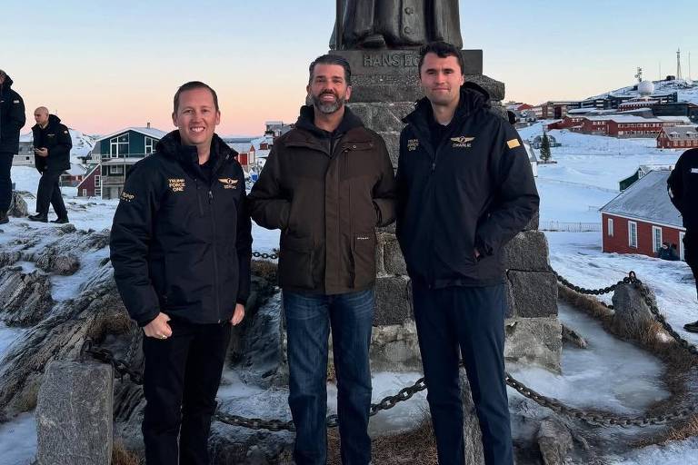 Três homens estão posando para a foto em frente a um monumento em uma área coberta de neve. O céu está claro， com tons de azul e laranja， indicando o início da manhã ou do final da tarde. Os homens estão vestidos com roupas de inverno， e há outras pessoas ao fundo， além de casas e uma paisagem montanhosa