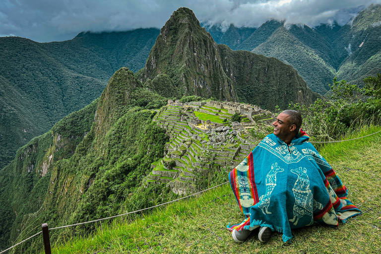 Um homem sentado em um gramado verde， usando um poncho colorido com estampas de lhamas， sorrindo enquanto observa a cidade inca de Machu Picchu ao fundo. A paisagem é montanhosa， com vegetação densa e nuvens cobrindo as montanhas. O céu está nublado， criando uma atmosfera dramática.