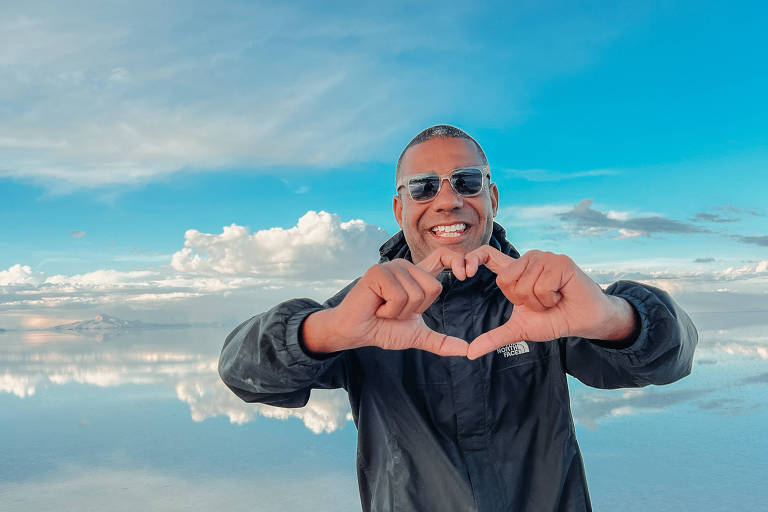 Um homem sorridente está em pé em uma superfície reflexiva， possivelmente um salar， com um céu azul e nuvens brancas ao fundo. Ele está usando óculos escuros e uma jaqueta preta， e faz um gesto de coração com as mãos.