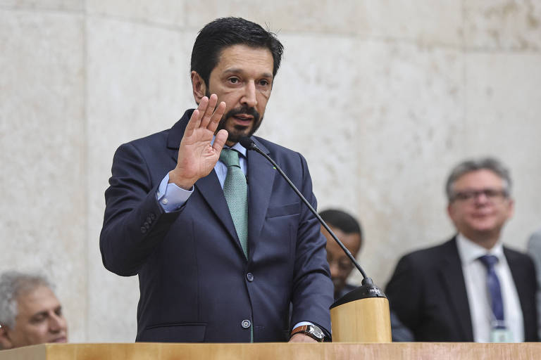 Um homem em um terno escuro e gravata verde está falando em um microfone， levantando a mão direita em um gesto de saudação ou ênfase. Ao fundo， outras pessoas estão assistindo， algumas sorrindo. O ambiente parece ser uma sala de conferências ou um plenário， com paredes claras.