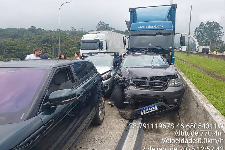 A imagem mostra um acidente de trânsito envolvendo vários veículos. Um caminhão azul está estacionado na parte de trás， enquanto um carro preto está colidido na lateral de outro veículo. A cena ocorre em uma estrada， com outros carros visíveis ao fundo. O céu está nublado， sugerindo um clima encoberto.