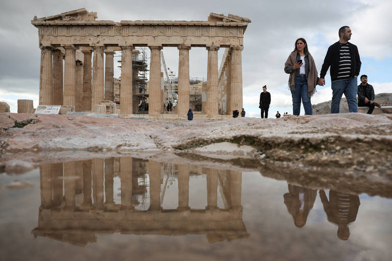 A imagem mostra o Partenon， um templo grego clássico， com colunas em pé e algumas partes em restauração. No primeiro plano， há uma poça de água refletindo a estrutura. Algumas pessoas estão caminhando em direção ao templo， enquanto outras estão paradas. O céu está nublado， sugerindo um clima sombrio.