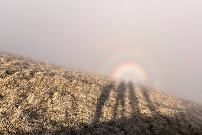 A imagem mostra uma colina coberta de grama seca， com sombras de pessoas projetadas no solo. Ao fundo， há um arco-íris visível， cercado por uma névoa que dá um aspecto etéreo à cena.