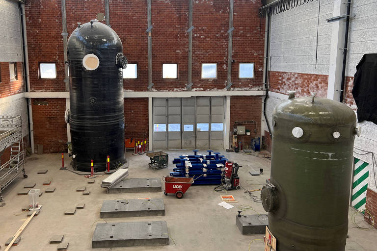 A imagem mostra o interior de um armazém industrial com paredes de tijolos expostos e janelas altas. No centro， há dois grandes tanques verticais， um preto e um verde. O chão está coberto com tábuas de madeira e há caixas e equipamentos espalhados， incluindo objetos azuis. A iluminação é natural， proveniente das janelas.