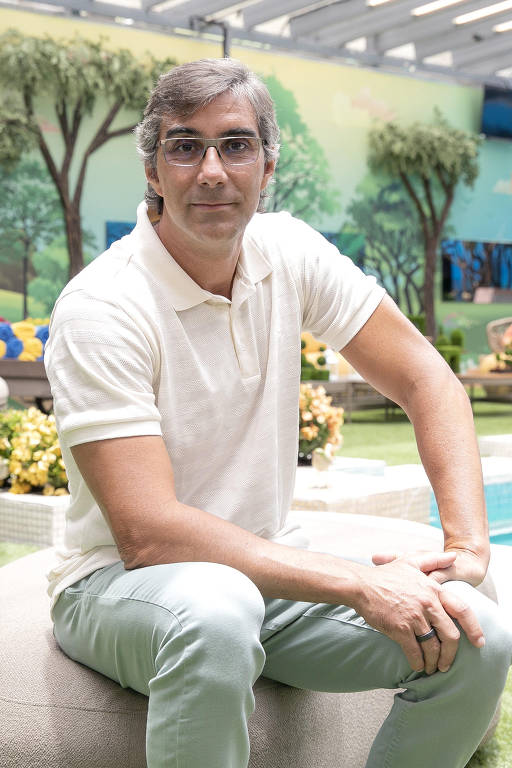 Um homem está sentado em um banco redondo em um ambiente externo. Ele usa uma camisa polo branca com listras e calças verdes claras. O fundo apresenta uma parede pintada com árvores e um gramado verde, além de flores coloridas e um espaço de lazer ao fundo.